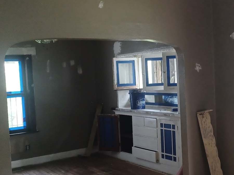 A room seen through an arch, with patched brown-gray walls, bare floorboards and a built-in cabinet with blue tape on its glass panes.