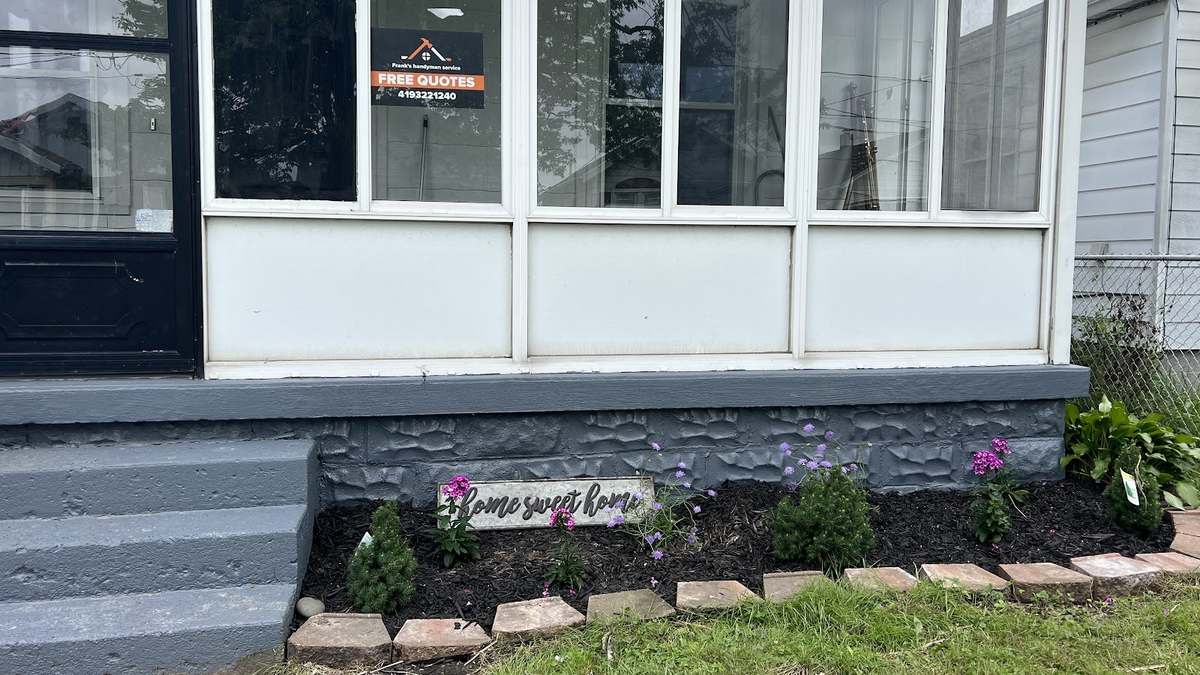 An enclosed white porch with a gray-painted foundation and steps, a Frank's Handyman Services free-quotes sign in the window, and a new flower bed with a Home Sweet Home sign in front.