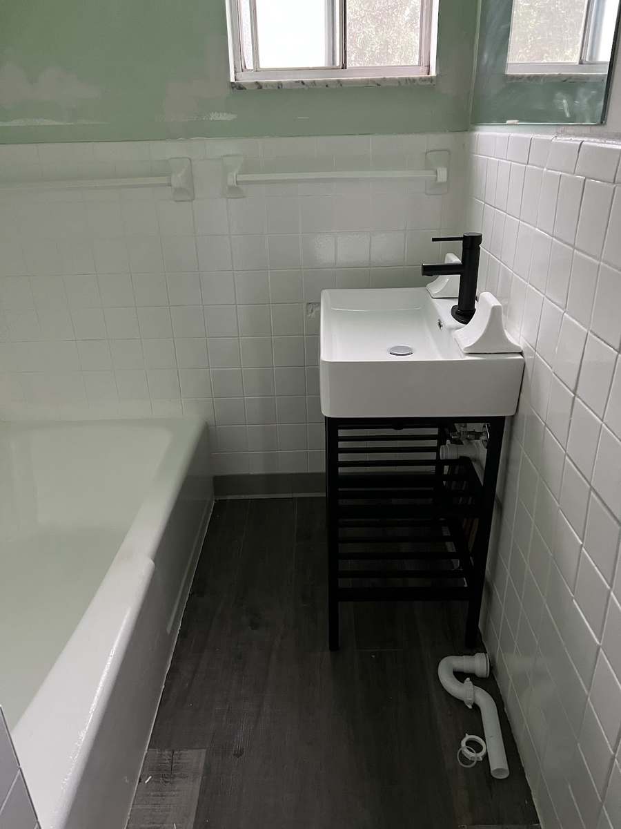A narrow bathroom with white wall tile, a white tub, a small white sink on a black stand with a black faucet, and dark plank flooring.