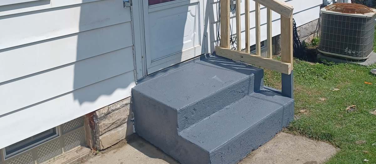 Gray-painted concrete steps with a new light-wood railing leading up to a white house's back door, with a green lawn beside them.